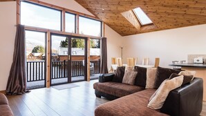 Un espacio habitable espacioso con un gran ventanal corredera que da a un balcón, una zona de estar acogedora con sofá y sillones, y una encimera de cocina con electrodomésticos.