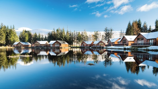 Una hilera de cabañas de madera junto a un lago tranquilo con reflejos, rodeadas de árboles y montañas nevadas.