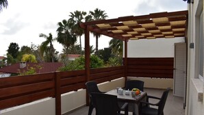 Une terrasse extérieure couverte, avec une table et des chaises, entourée d’une clôture en bois et de palmiers.