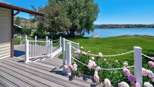 A wooden deck with white railings and a chain barrier leading to a grassy area with flowers and a lake view. - Lakefront single story house on Hauser Lake (Helena)