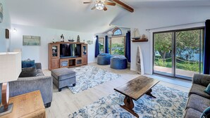 A spacious living room with a large TV, comfortable seating, a wooden coffee table, and a view of the outdoors through large windows. - Lakefront single story house on Hauser Lake (Helena)