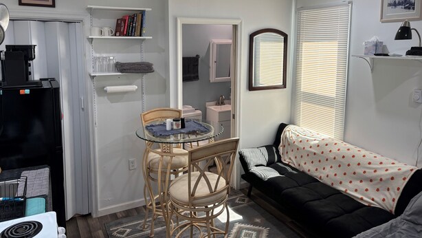Ein Wohnzimmer mit einer schwarzen Couch, einem Glastisch mit Stühlen, einer Küchenzeile mit Kühlschrank und einem Deckenventilator.