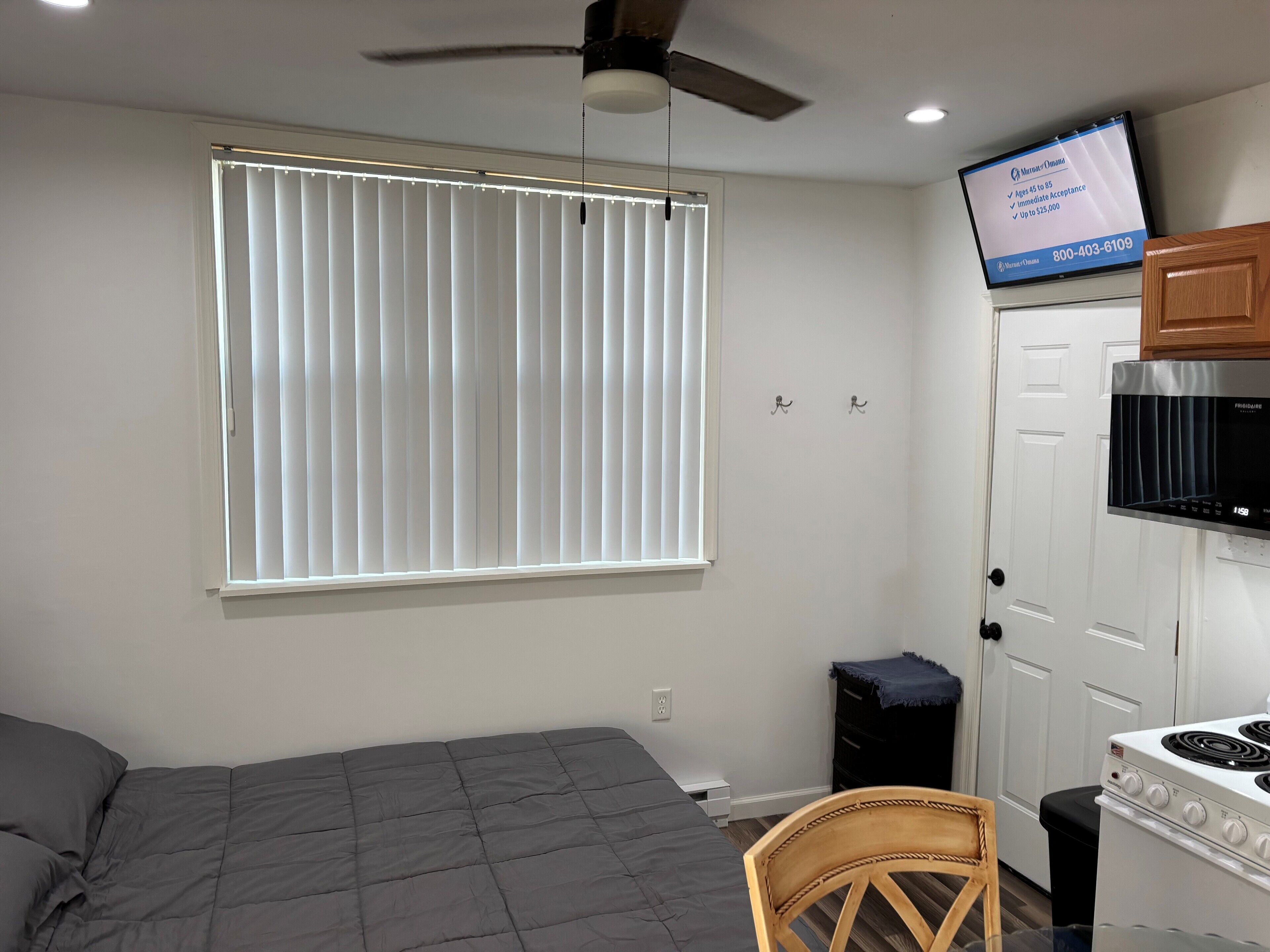 A bedroom with a bed, a ceiling fan, a window with blinds, a TV mounted on the wall, and a kitchen area with a stove and cabinets.
