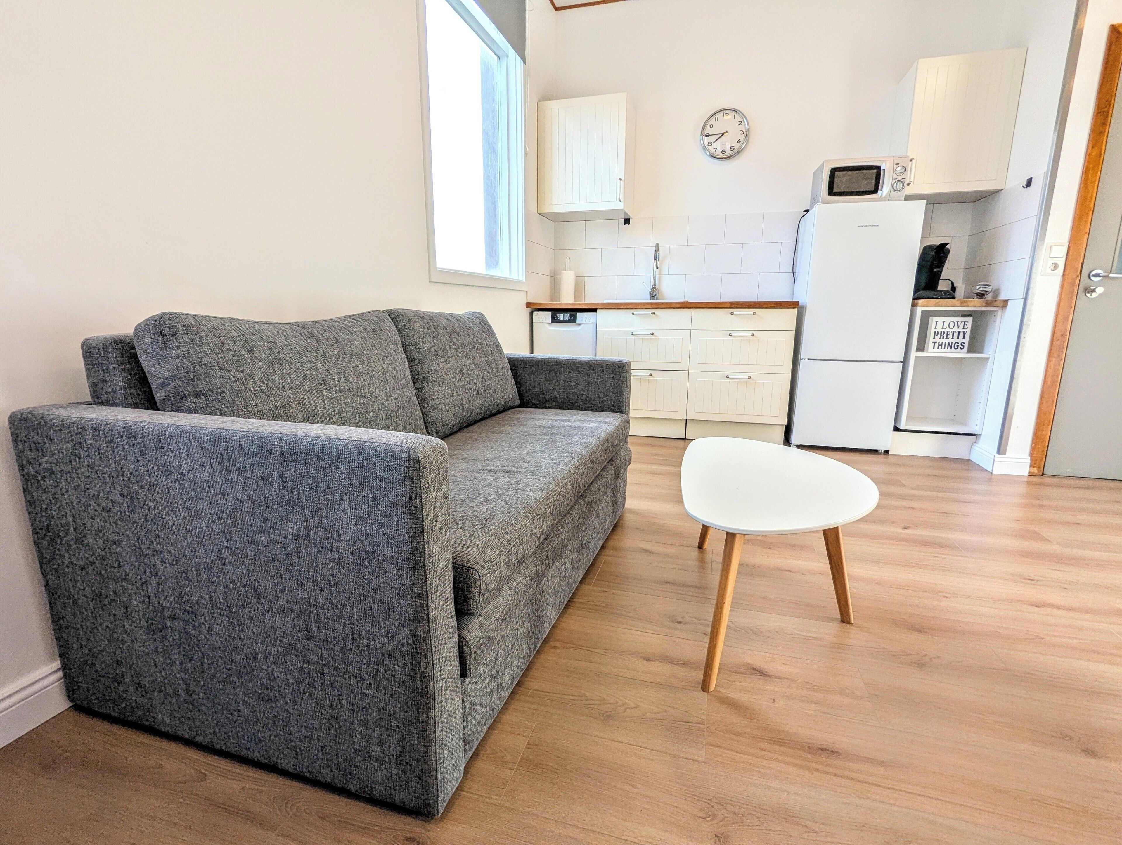 A living room with a grey sofa, a white table, wooden flooring, a kitchen with white cabinets, a microwave, and a clock on the wall.
