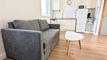 A living room with a grey sofa, a white table, wooden flooring, a kitchen with white cabinets, a microwave, and a clock on the wall.