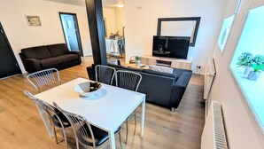 A modern living room with a dining area, a sofa, a TV, and a window with plants. - Central Apartments  (Vestmannaeyjar)