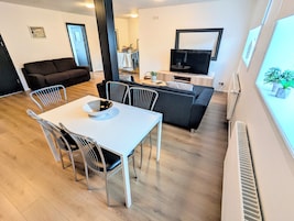 Una sala de estar moderna con comedor, sofá, televisor y ventana con plantas.