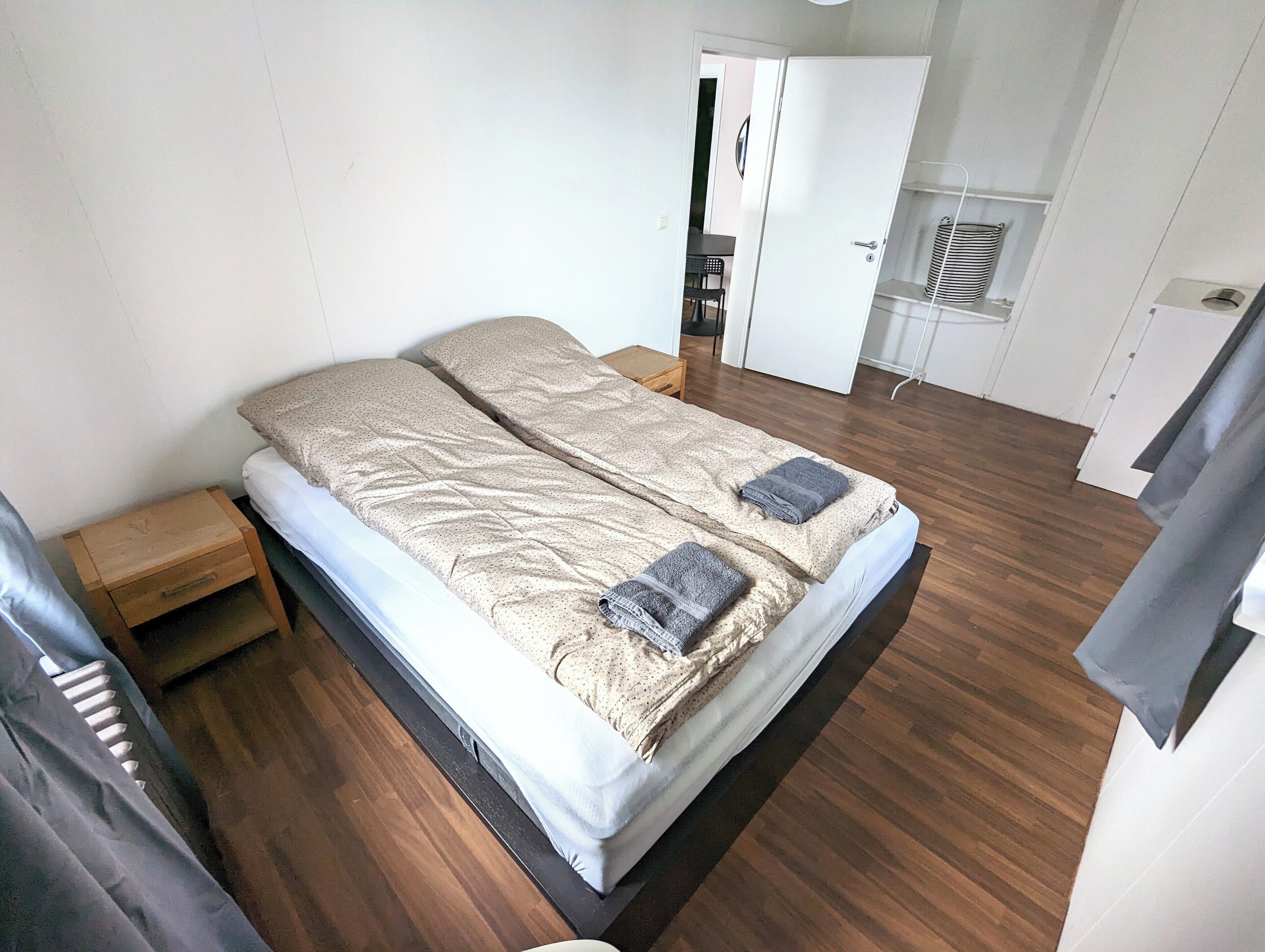 Un lit double avec une couvre-lit à motifs et deux serviettes pliées, une table de chevet en bois, une petite table et une armoire blanche dans une pièce dont le sol est recouvert de parquet.