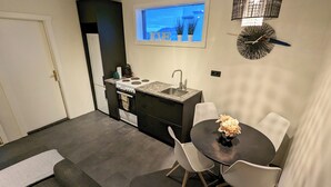 A compact kitchen with a built-in stove, sink, and window with a view. There is a dining area with a table and chairs. - Central Apartments  (Vestmannaeyjar)
