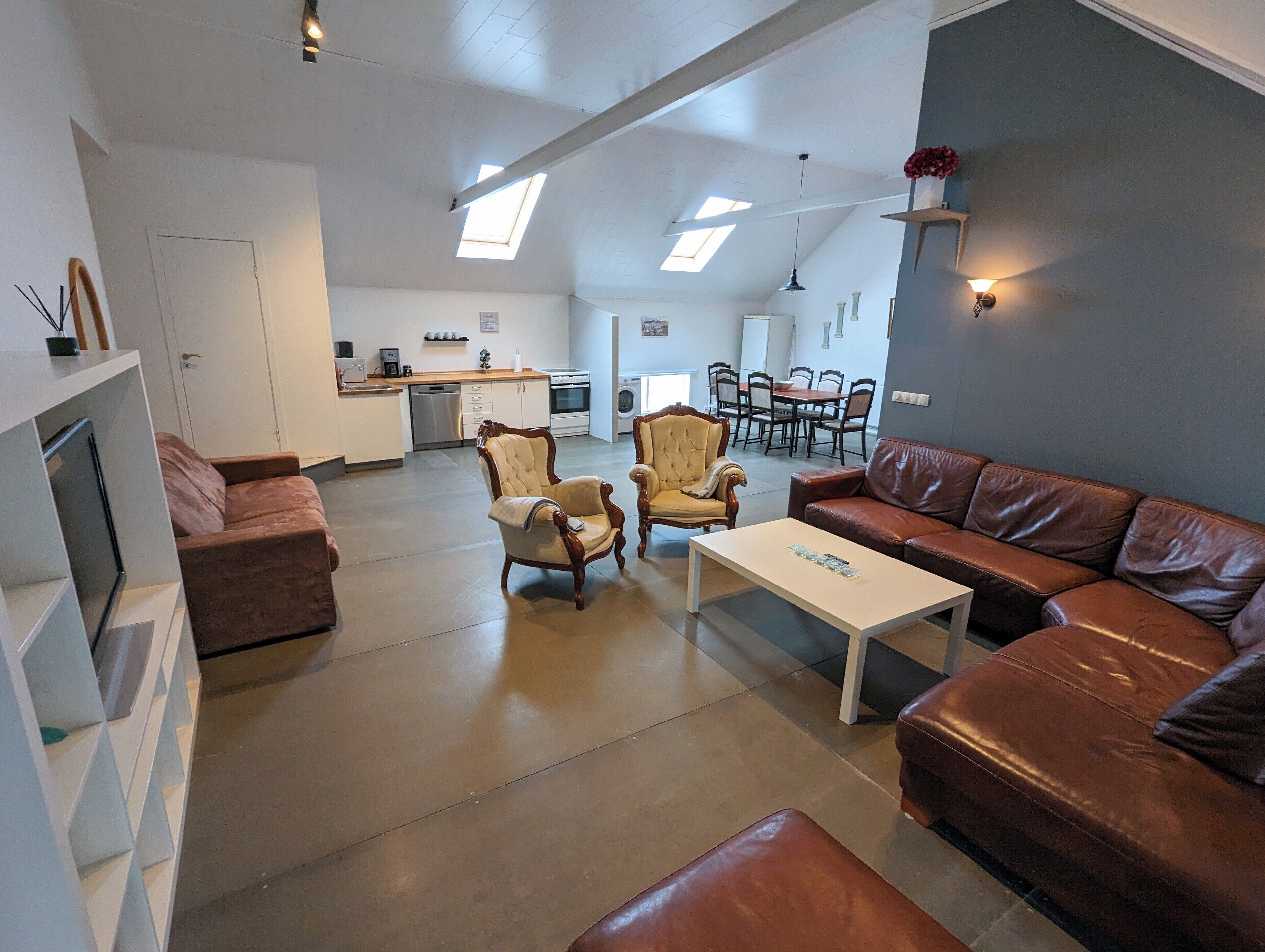 A spacious living area with a brown leather sofa, a white coffee table, and a beige armchair. A kitchen with a dishwasher and a dining area with a table and chairs are visible in the background.