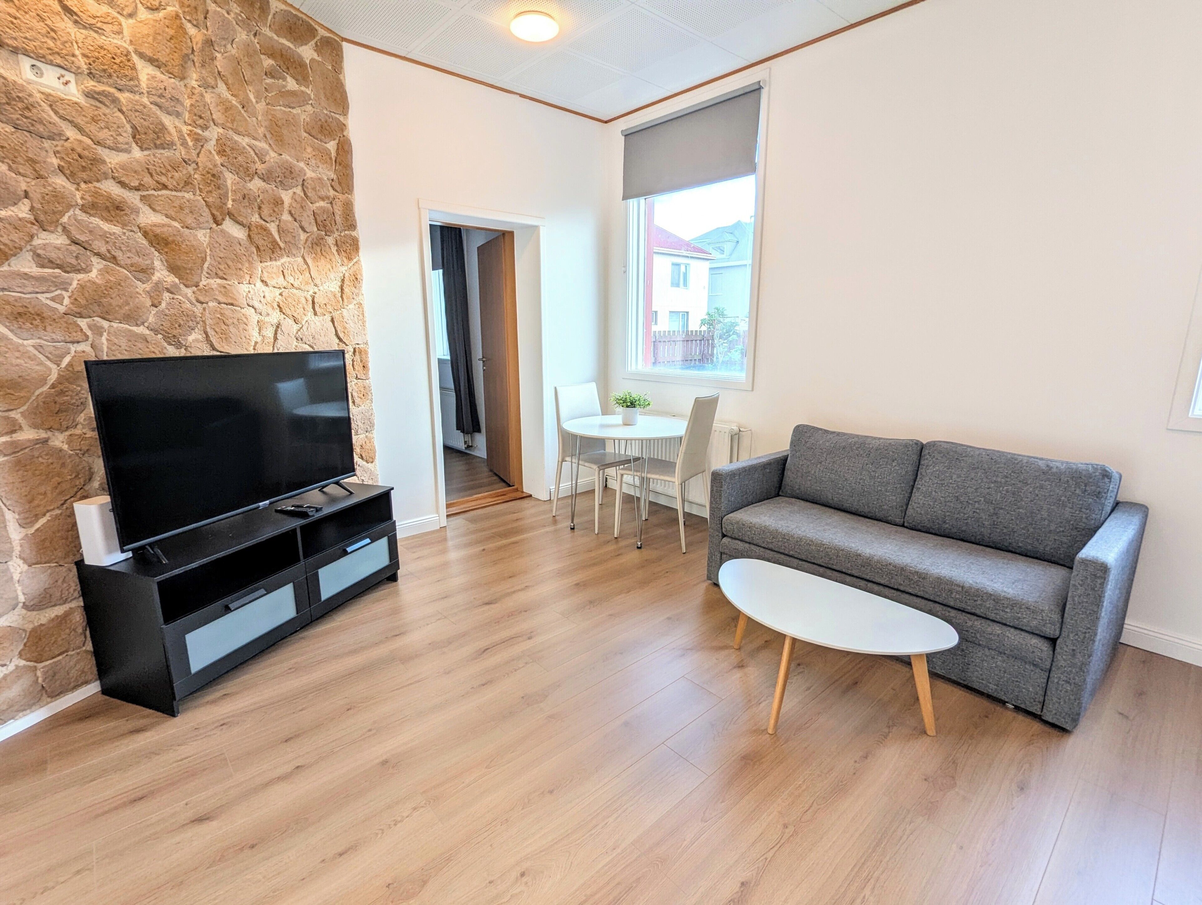 Una sala de estar con pared de piedra, un televisor de pantalla plana, un sofá gris, una pequeña mesa blanca y un comedor con mesa y sillas blancas.