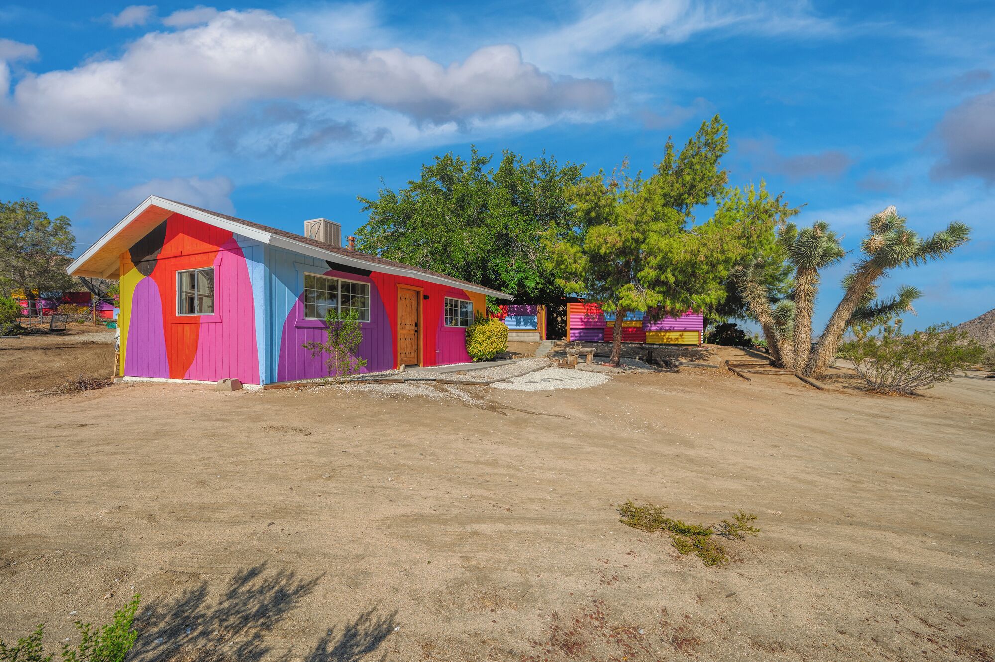 A colorful, single-story house with a sloped roof, surrounded by desert vegetation and clear skies.