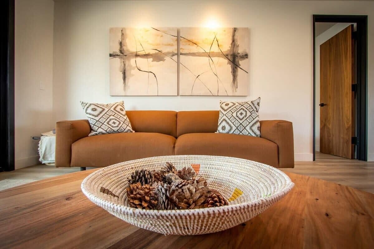 A living room with a brown sofa, two patterned throw pillows, a woven basket on a wooden table, and a large abstract painting on the wall.