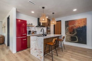 A modern kitchen with a red refrigerator, marble island, wooden bar stools, pendant lights, and a large abstract painting on the wall. - @ Marbella Lane - The Wine Mine Desert Escape (Joshua Tree)