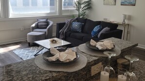 A modern living room with a dark grey sofa, a glass coffee table, and a large window with blinds. - 4 Downtown Indy SkyWalk to Gainbridge Lucas Oil Conv Cntr (Indianapolis)