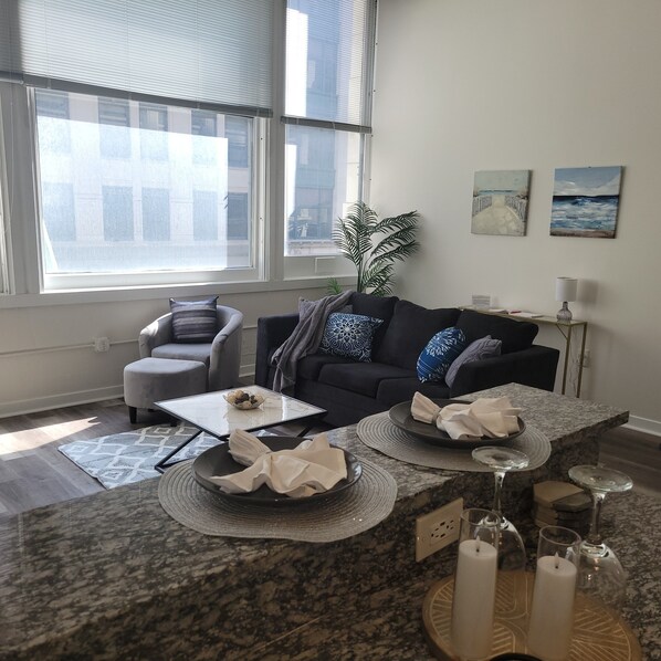A modern living room with a dark grey sofa, a glass coffee table, and a large window with blinds. - #4 Downtown Free Parking- Walk to Lucas Oil, Conv Center, Gainbridge (Indianapolis)
