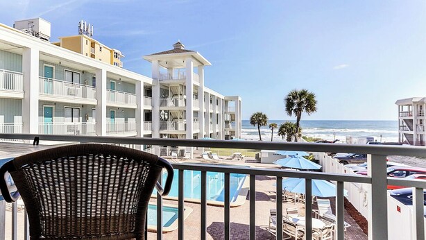 A balcony with a wicker chair, a pool area with umbrellas, and a view of a beach and ocean.