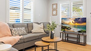 A living room with a grey sofa, two round wooden tables, a TV on a stand, and a window with blinds. - @Marbella Lane - Serene Haven Retreat (Ontario)