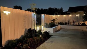 A well-lit outdoor area with a wooden fence, a concrete pathway, and various plants and shrubs. - @Marbella Lane - Serene Haven Retreat (Ontario)