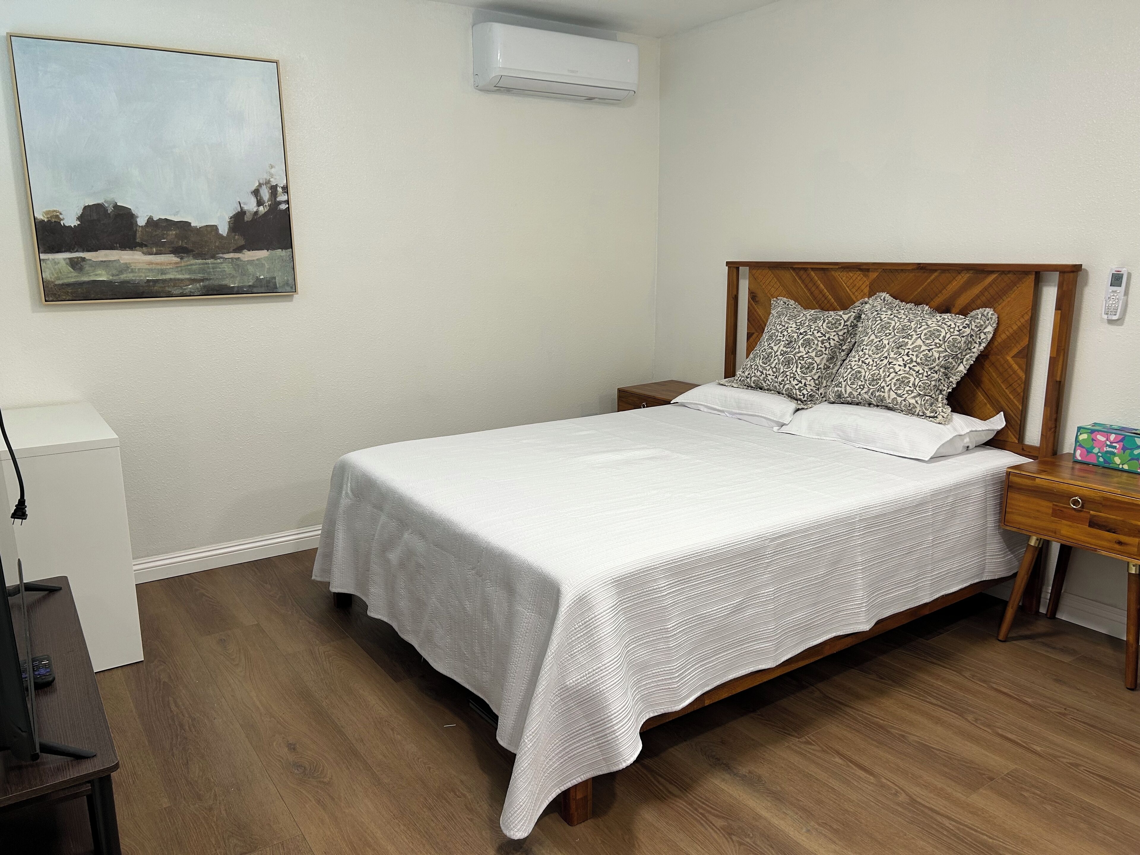 A neatly made bed with a white bedspread and two patterned pillows, a wooden nightstand with a box of tissues, and a painting on the wall.