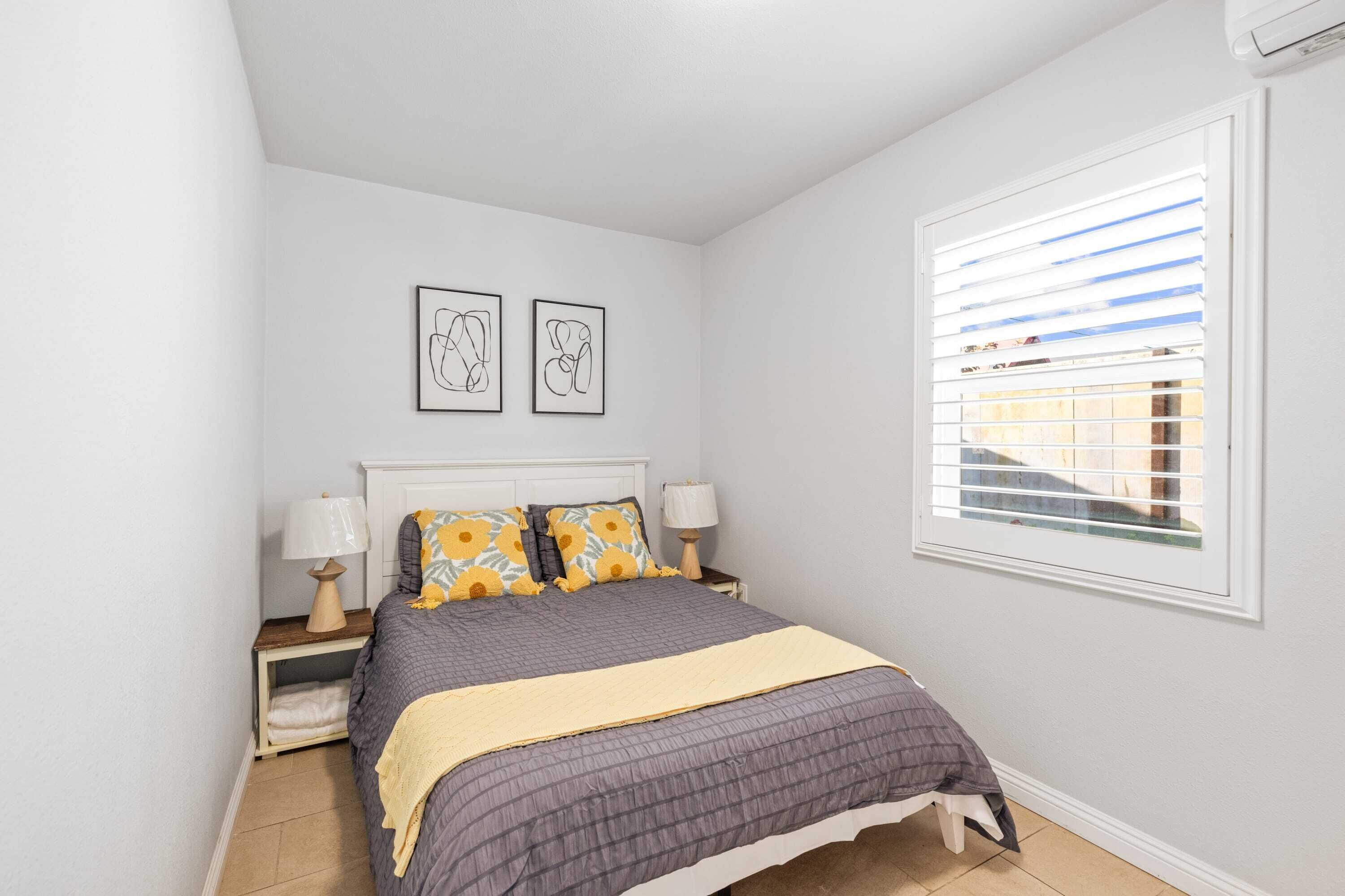 A bedroom with a bed, two bedside tables with lamps, a window with blinds, and two framed pictures on the wall.