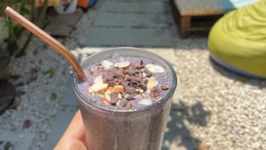 Une main tenant un verre de smoothie avec une paille, une plante verte et un pouf jaune en arrière-plan.
