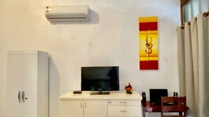 A room with a flat-screen TV on a white cabinet, a small table with a laptop, a speaker, and a decorative item.