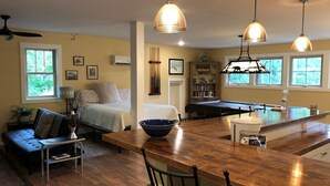 Ein gemütliches Wohnzimmer mit einer Couch, einem Esstisch und einer Küchenzeile mit Bar. Mehrere Fenster sorgen für natürliches Licht.