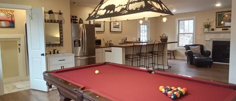 A pool table in a room with a kitchen area, a bar, and a fireplace.