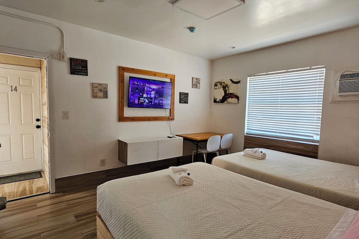 A hotel room with two beds, a desk, a TV, and a window with blinds.