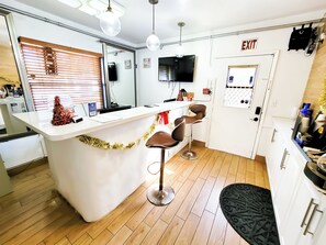 A modern kitchen with a white countertop, two bar stools, a television, and an exit sign. - Stay Inn Boutique Hotel  (Dania Beach)