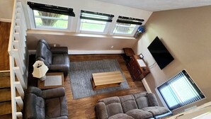 A spacious living area with a sectional sofa, a coffee table, and a TV on the wall. There are three windows with blinds, a staircase, and a wooden floor. - Returning to the summer rental market fully renovated 4 Br/2 bath home A/C (Narragansett)