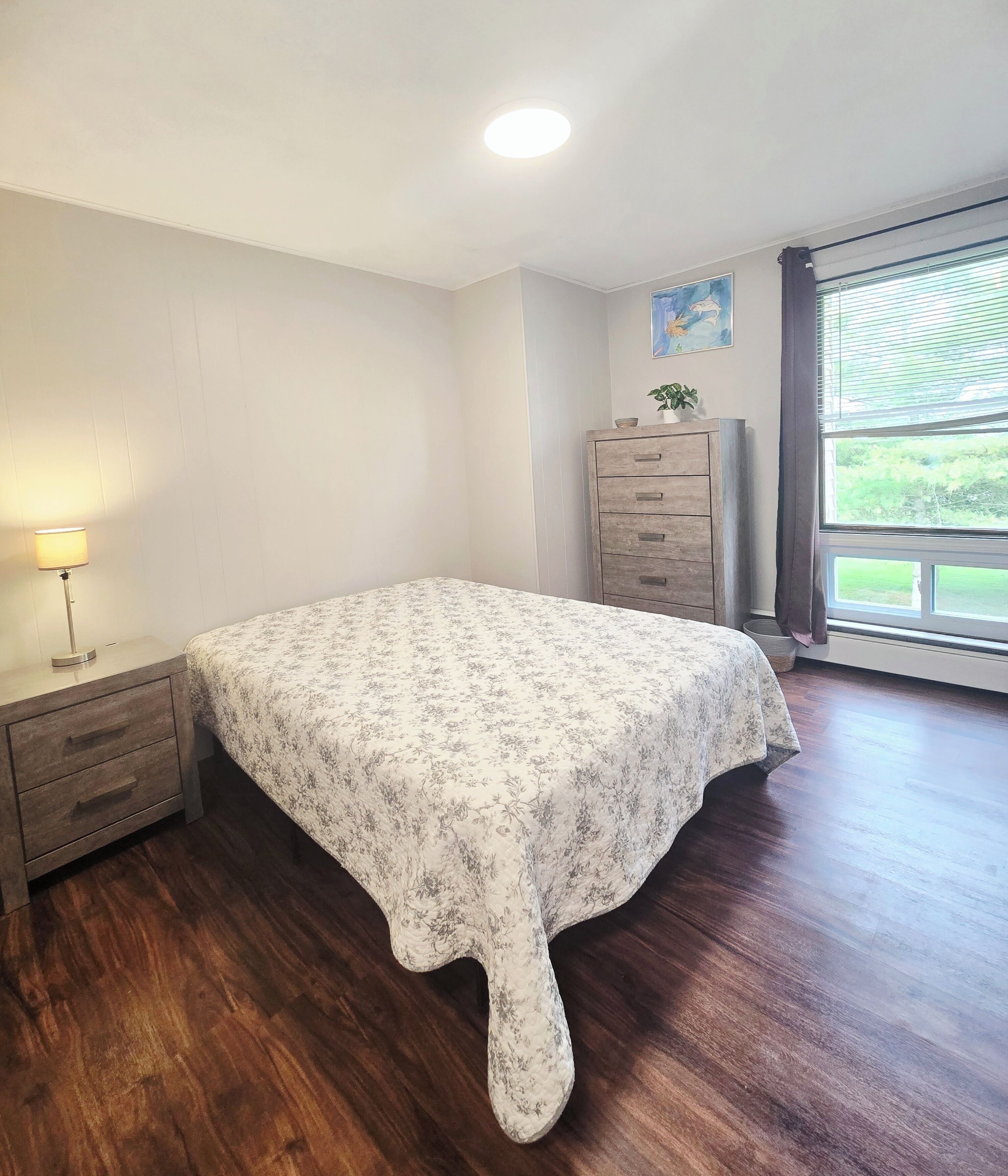 Ein Schlafzimmer mit Blumenmuster-Bettdecke, einem Holzkleiderschrank, einem Nachttisch mit Lampe und einem Fenster mit Blick auf Grünflächen.
