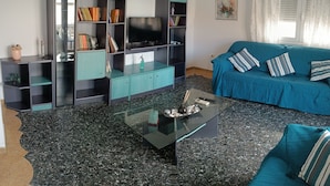 A living room with a teal sofa, a glass coffee table, and a black entertainment unit with books and a TV. - PAP'S HOUSE  (Rodes)