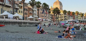 A beach with people sunbathing and walking, a large dome building in the background, and colorful buildings along the promenade.