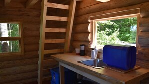 A wooden cabin with a kitchen area featuring a sink, a window with a view of greenery, and a ladder leading to an upper level.