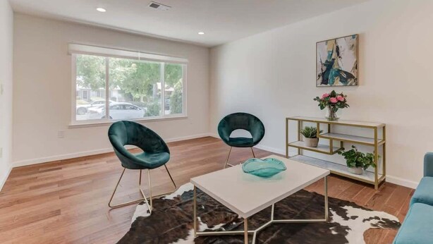 Een moderne woonkamer met twee teal fauteuils, een witte salontafel, een gouden console met planten en een groot raam met uitzicht op een parkeerplaats.