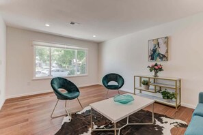 Una sala moderna con dos sillones color turquesa, una mesa de centro blanca, una consola dorada con plantas, y un ventanal con vista a un estacionamiento.