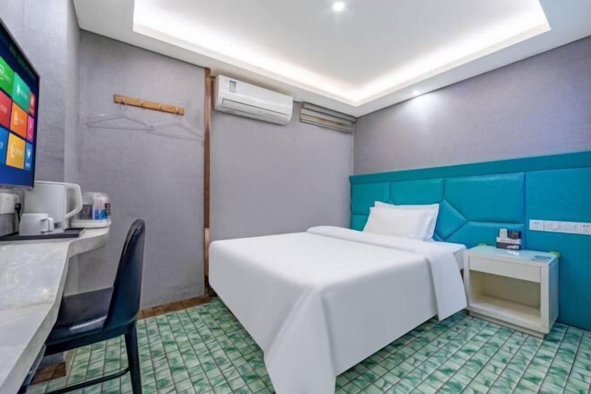 A hotel room with a bed, a desk, a chair, a nightstand, and an air conditioning unit.