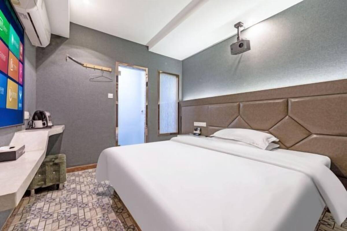 A hotel room with a bed, a desk with a coffee maker, a TV, and a wall-mounted projector.