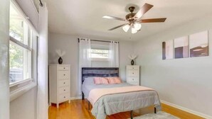 Un dormitorio con cama, ventilador de techo, cómoda, mesita de noche y ventana con cortinas.