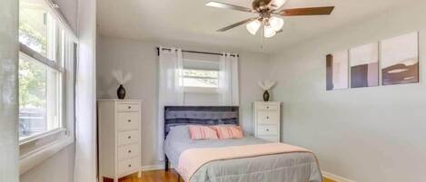 Un dormitorio con cama, ventilador de techo, cómoda, mesita de noche y ventana con cortinas.