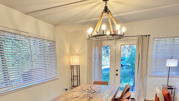 A dining room with a long wooden table, cushioned chairs, a chandelier, and large windows with blinds.