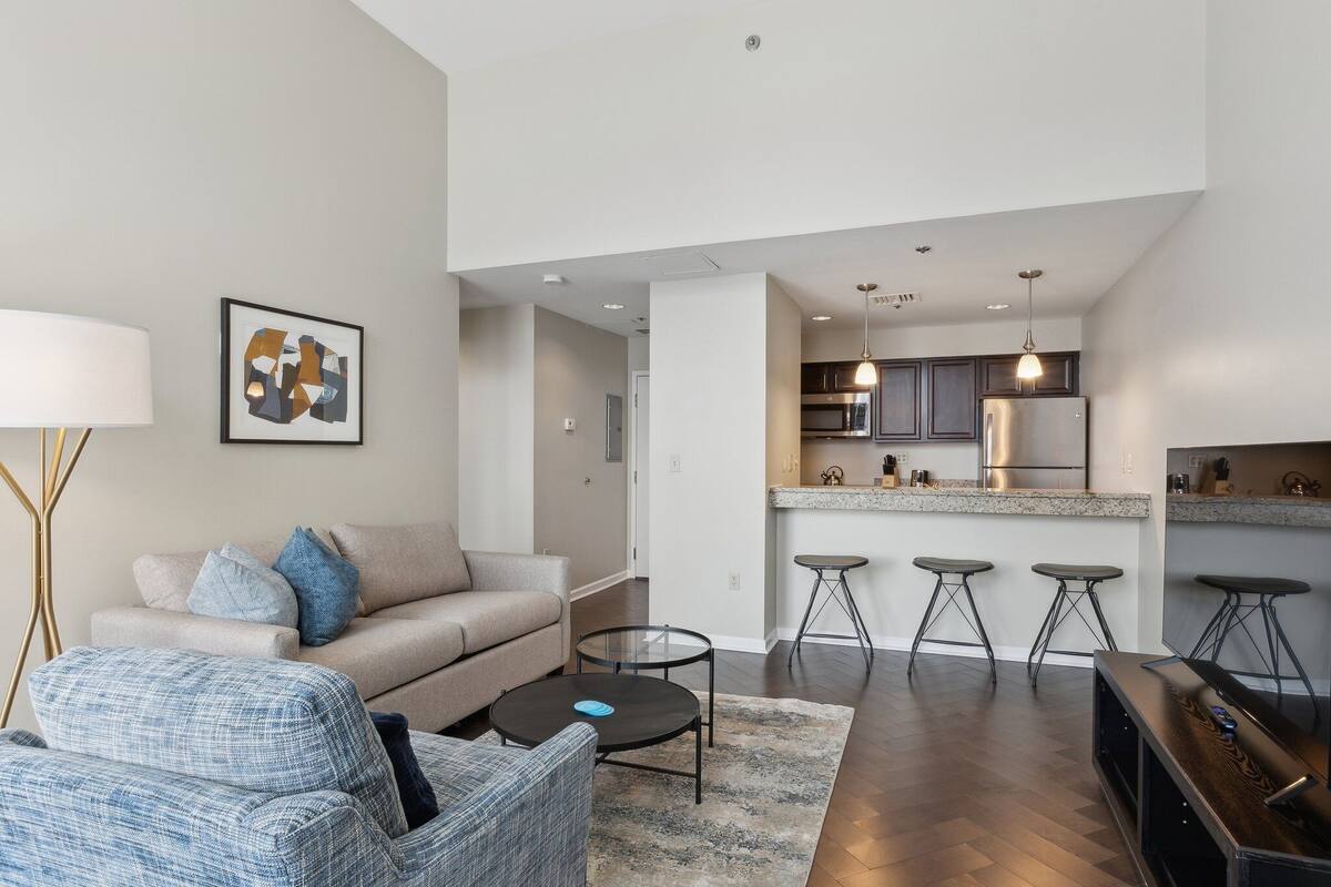 Una sala de estar moderna con un sofá, una mesa de centro y un televisor. Se ve una cocina con una barra y taburetes al fondo.