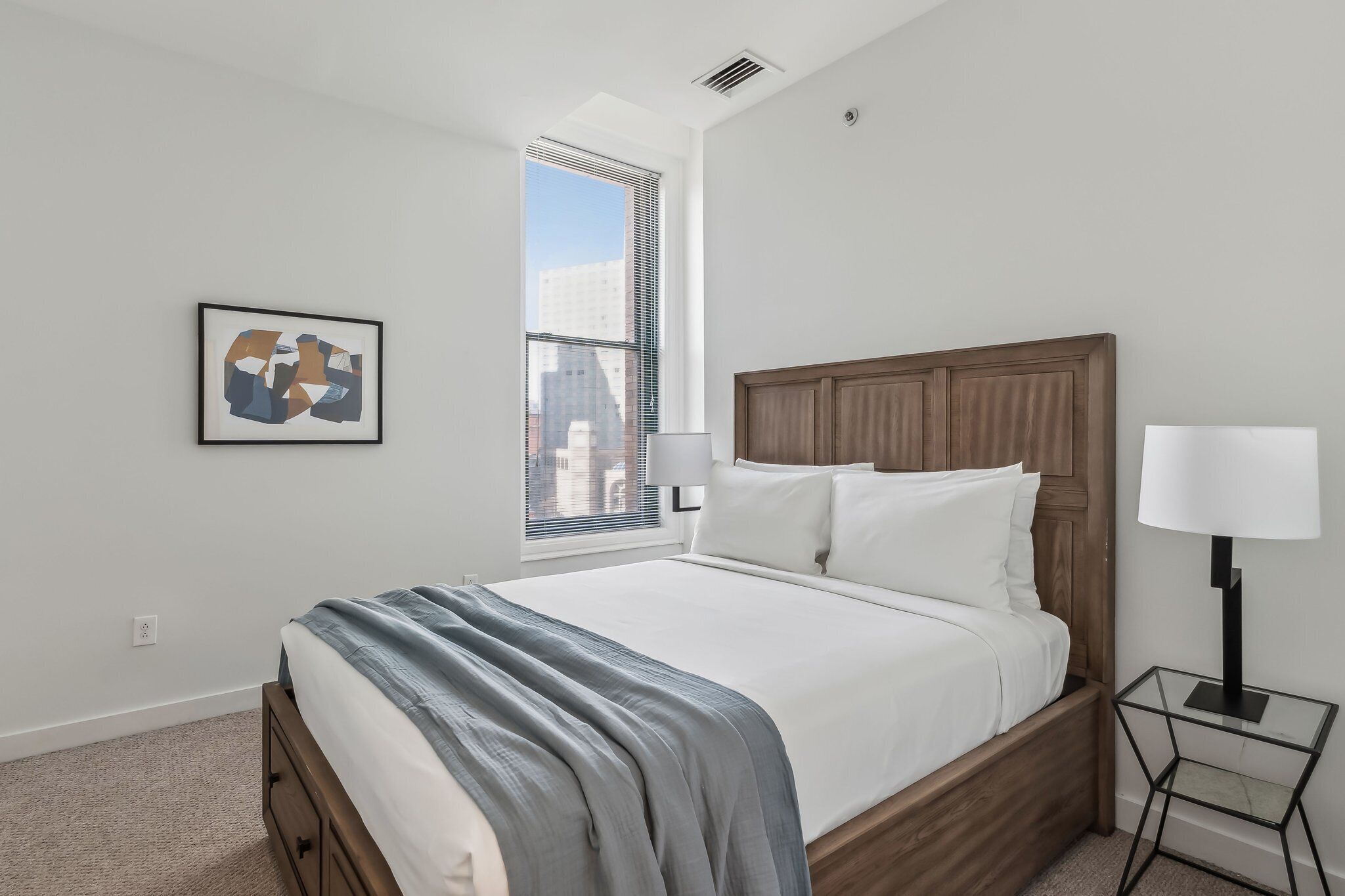 Un dormitorio moderno con cabecera de madera, una cama grande, un cuadro abstracto enmarcado, una mesita de noche con lámpara y vistas a la ciudad a través de la ventana.
