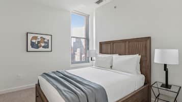 A modern bedroom with a wooden headboard, a large bed, a framed abstract painting, a bedside table with a lamp, and a city view through the window.