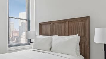 A neatly made bed with white linens in a room with a wooden headboard, a bedside lamp, and a view of a cityscape through a window with blinds.