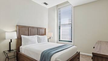 A neatly made bed with a wooden headboard, a bedside table with a lamp, and a window with blinds.