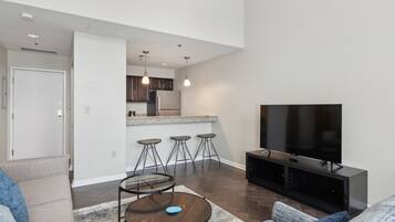 Een moderne woonkamer met een bank, salontafel en een televisie. Er is een keuken met een koelkast, barstoelen en een aanrecht.