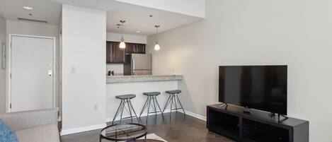 A modern living room with a sofa, coffee table, and a TV. There is a kitchen area with a refrigerator, bar stools, and a countertop.